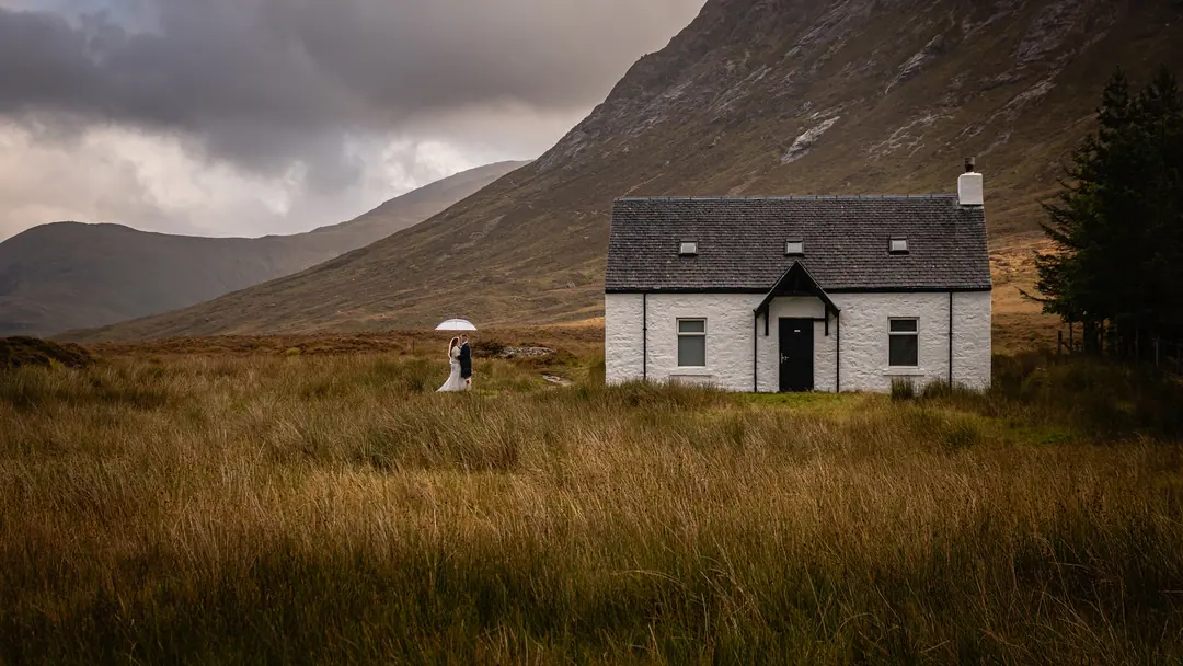 Couple beneath a white umbrella beside a whitewashed cottage in Glencoe