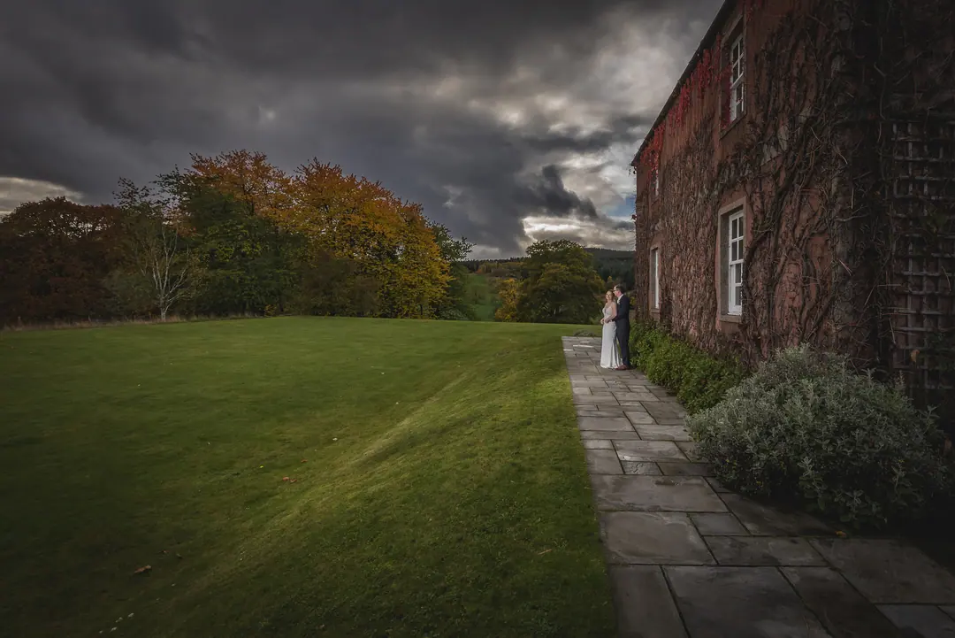 Couple on a lawn beside a red-brick country house beneath a brooding sky