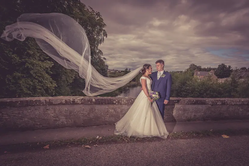 Veil streaming in the wind as a couple stand on an old stone bridge