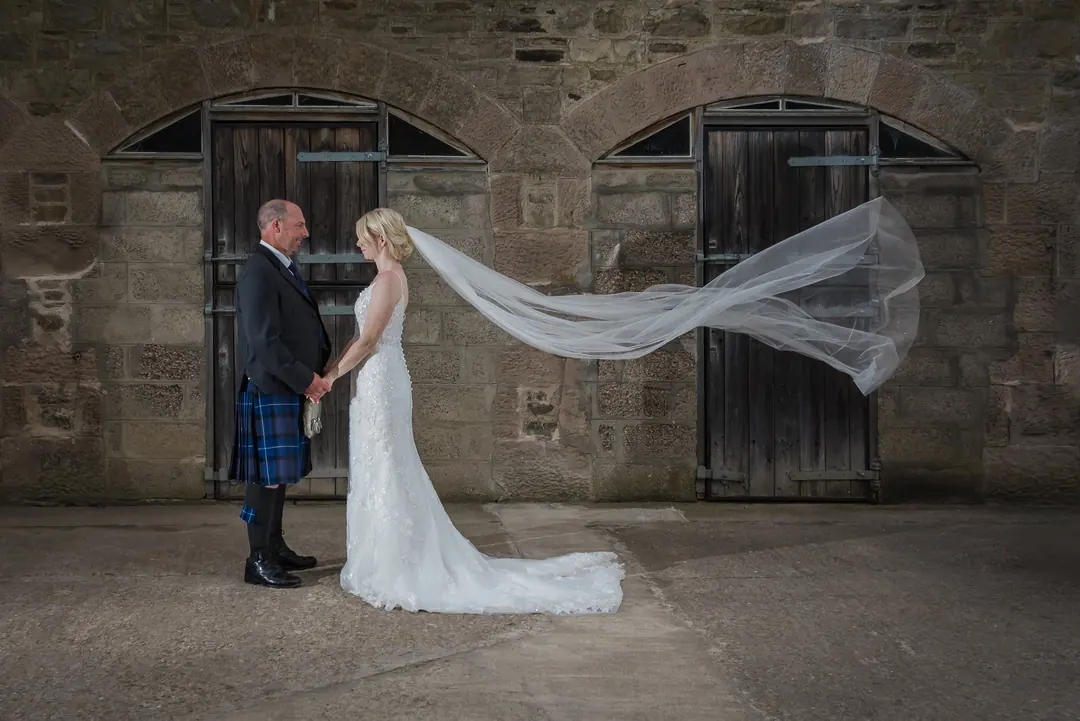 Groom in a kilt holding his bride’s hands as her veil flies in the wind against old stable doors