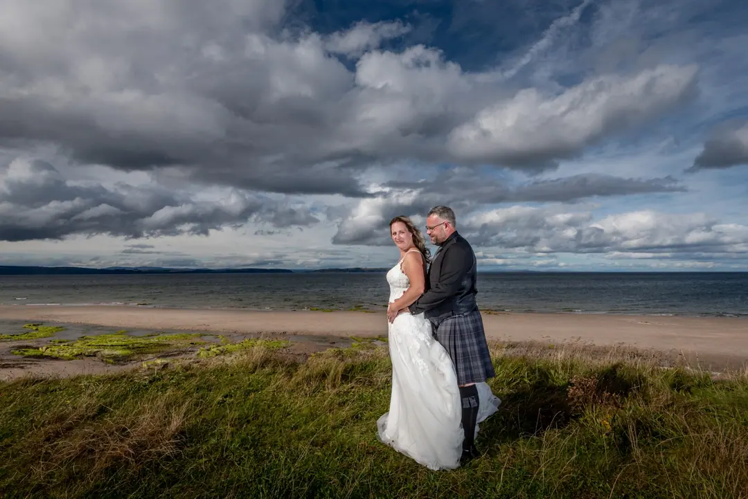 Bride and groom on the grassy dunes above a beach beneath dramatic clouds